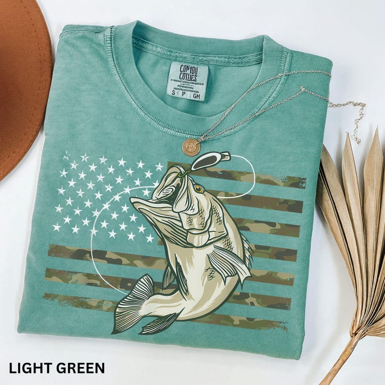 May include: Light green t-shirt featuring a graphic of a fish with a fishing lure, set against a camouflage and star-spangled banner design. The shirt has a crew neck and short sleeves. The text "LIGHT GREEN" is at the bottom of the image.
