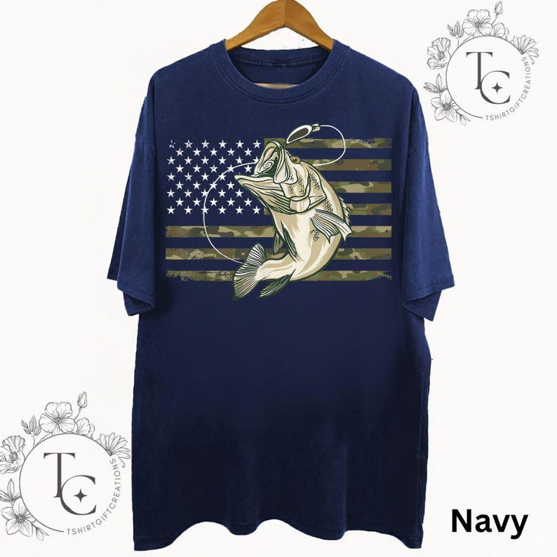 May include: Navy blue t-shirt featuring a graphic of a fish jumping out of the water, with a fishing hook and line. The design incorporates the American flag with a camouflage pattern. The word "Navy" is printed at the bottom right.