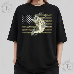 May include: Black crew neck t-shirt featuring a graphic of a fish jumping out of the water with a fishing hook. The background is an American flag with a camouflage pattern.