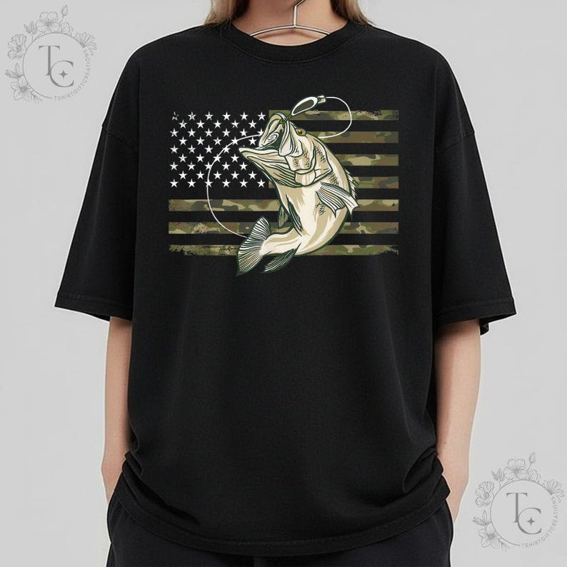 May include: Black crew neck t-shirt featuring a graphic of a fish jumping out of the water with a fishing hook. The background is an American flag with a camouflage pattern.