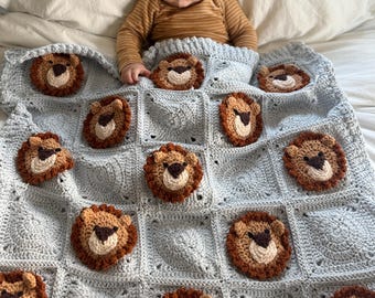 May include: A light blue crocheted baby blanket featuring multiple lion faces. The lion faces are brown, tan, and cream-colored, with dark brown noses. The blanket is square-patterned and appears soft and cozy.