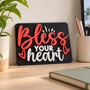 May include: A rectangular black sign featuring the raised 3D text "Bless YOUR heart" in red and white lettering. The word "Bless" is in red, "YOUR" is in white, and "heart" is in white. Two red heart icons are positioned at the corners.