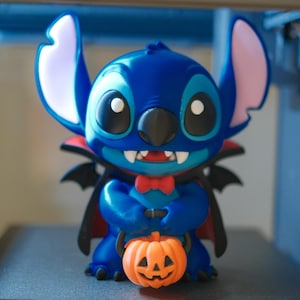May include: A blue Stitch figurine dressed as a vampire with a black cape featuring a red interior lining, a red bowtie, and large ears with pink inner sections. The character holds a small orange jack-o'-lantern pumpkin bucket.