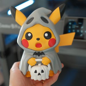 Może przedstawiać: Żółta figurka Pikachu w szarym kostiumie ducha z kapturem, czarnymi końcówkami uszu i czerwonymi policzkami. Postać trzyma małe białe wiaderko w kształcie dyni z czarną twarzą dyni i czarnym emblematem nietoperza na piersi.