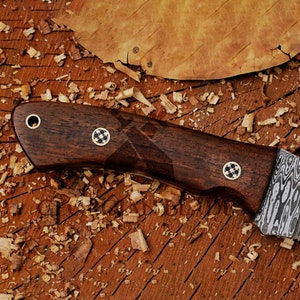 May include: A close-up of a dark brown wooden knife handle with a decorative pattern and two metal accents. The blade is made of a patterned metal.