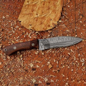 May include: A close-up of a knife with a wooden handle and a Damascus steel blade. The knife is lying on a wooden surface with wood shavings.
