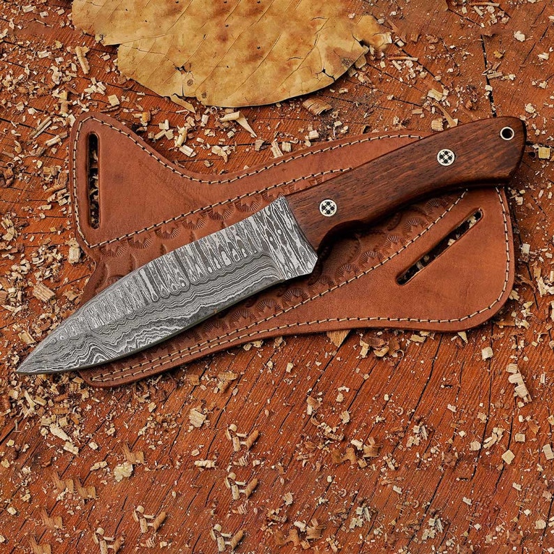 May include: A brown leather sheath with a decorative pattern holds a knife with a Damascus steel blade and a dark wood handle.