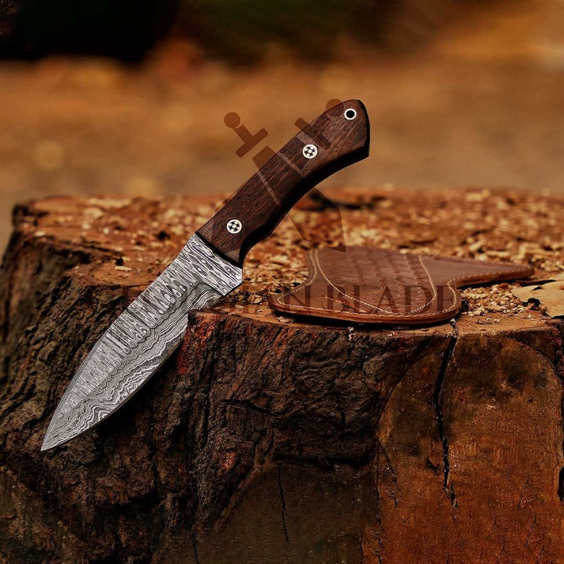 May include: A Damascus steel hunting knife with a wooden handle and a leather sheath. The knife has a patterned blade and is resting on a tree stump.