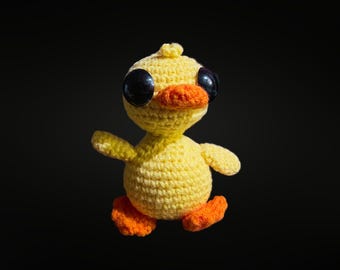 May include: A handmade yellow crochet duck with large black eyes and an orange beak and feet. The duck has a rounded body and small wings, giving it a whimsical appearance. The background is a solid black.