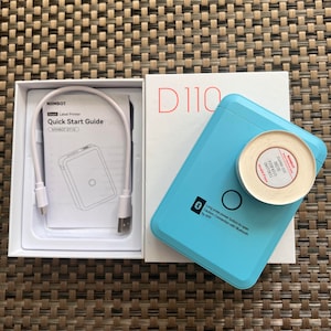 May include: A blue and white portable label printer with a roll of white label tape. The printer is labelled "NIMBOT D110" and has a Bluetooth symbol. The packaging includes a quick start guide and a USB cable.