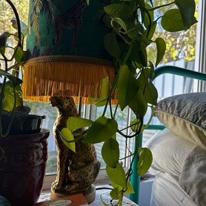 May include: A decorative table lamp with a green lampshade featuring a giraffe and palm tree design. The lamp has a gold fringe and sits atop a gold leopard statue. A book titled "The Secret History" is visible.