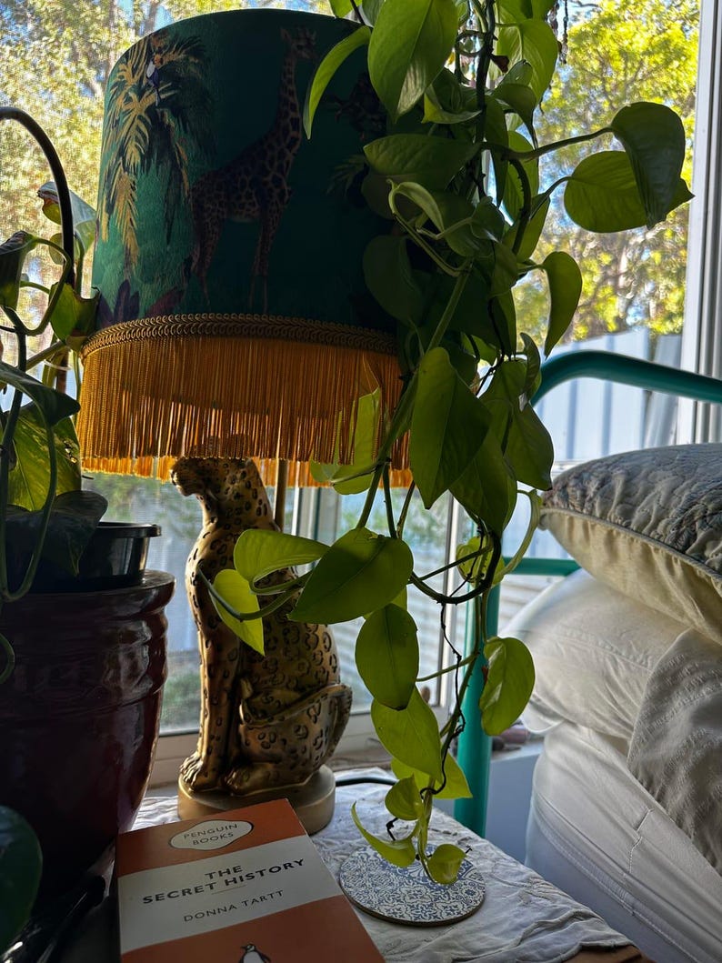 May include: A decorative table lamp with a green lampshade featuring a giraffe and palm tree design. The lamp has a gold fringe and sits atop a gold leopard statue. A book titled "The Secret History" is visible.