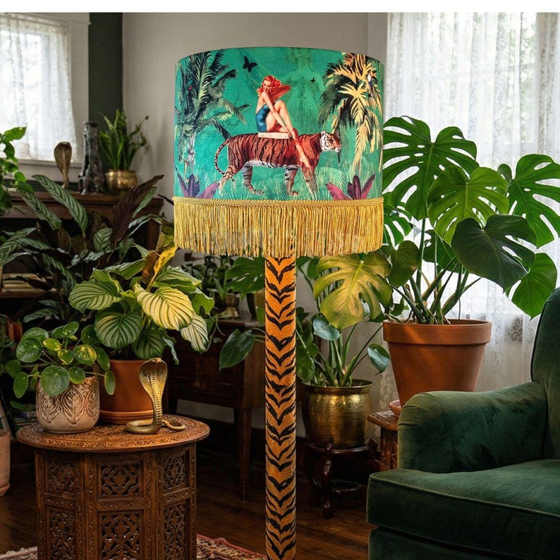 May include: A floor lamp with a vibrant, jungle-themed lampshade featuring a woman riding a tiger. The shade has a green background with palm trees and a gold fringe. The lamp's base has a tiger stripe pattern.