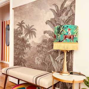 May include: A room featuring a large sepia-toned jungle mural, a bench with dark stripes, and a table lamp with a green lampshade. The lampshade has a jungle scene with a tiger and fringe. A small side table and colorful pillows complete the decor.