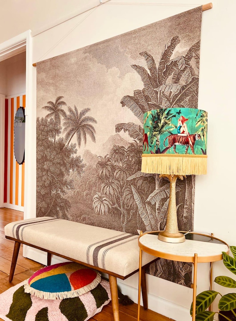 May include: A room featuring a large sepia-toned jungle mural, a bench with dark stripes, and a table lamp with a green lampshade. The lampshade has a jungle scene with a tiger and fringe. A small side table and colorful pillows complete the decor.