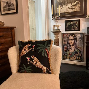 May include: A white armchair with a black and gold throw pillow featuring a cheetah print and palm trees. The pillow has a fringe trim. The chair is in a living room with artwork on the walls.