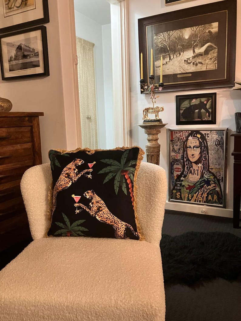 May include: A white armchair with a black and gold throw pillow featuring a cheetah print and palm trees. The pillow has a fringe trim. The chair is in a living room with artwork on the walls.