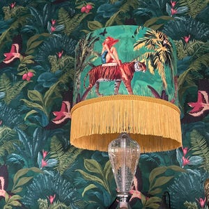 May include: A decorative table lamp with a clear glass base and a vibrant green lampshade. The lampshade features a woman riding a tiger in a jungle scene, with a gold fringe trim. The background is a dark green jungle wallpaper.