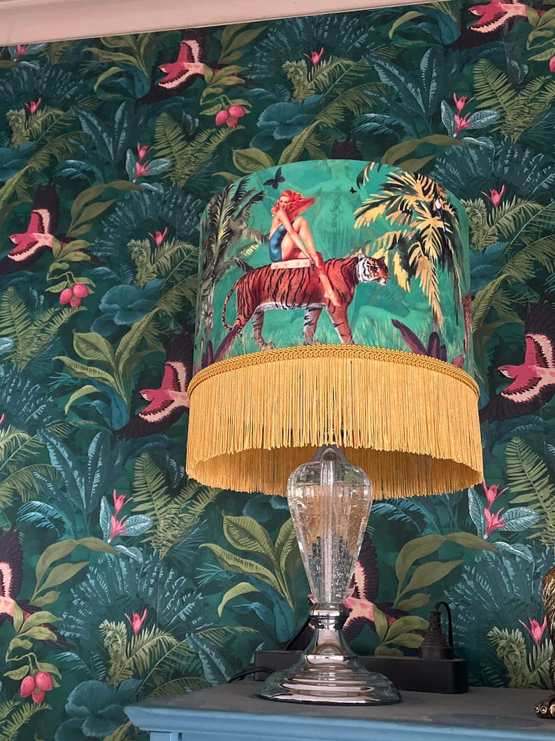 May include: A decorative table lamp with a clear glass base and a vibrant green lampshade. The lampshade features a woman riding a tiger in a jungle scene, with a gold fringe trim. The background is a dark green jungle wallpaper.