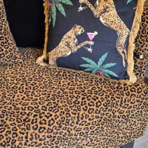May include: A black and gold throw pillow with a fringe border features a cheetah print design with palm trees and cocktails. The pillow is on a leopard print sofa.