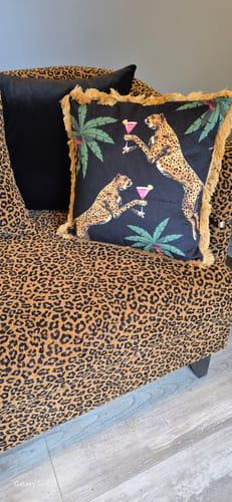 May include: A black and gold throw pillow with a fringe border features a cheetah print design with palm trees and cocktails. The pillow is on a leopard print sofa.