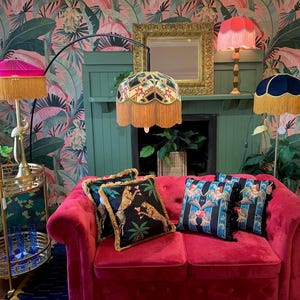 May include: A pink velvet sofa with gold trim, three lamps with fringed shades, and two throw pillows with animal and floral patterns. The lamps are on a gold stand and a fireplace mantel. The wallpaper is a tropical print with pink and green leaves.