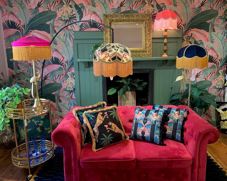 May include: A pink velvet sofa with gold trim, three lamps with fringed shades, and two throw pillows with animal and floral patterns. The lamps are on a gold stand and a fireplace mantel. The wallpaper is a tropical print with pink and green leaves.