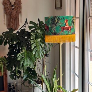May include: A floor lamp with a green lampshade featuring a jungle scene with a tiger and a woman. The lampshade has a yellow fringe at the bottom. The lamp is gold-colored and stands among various houseplants.