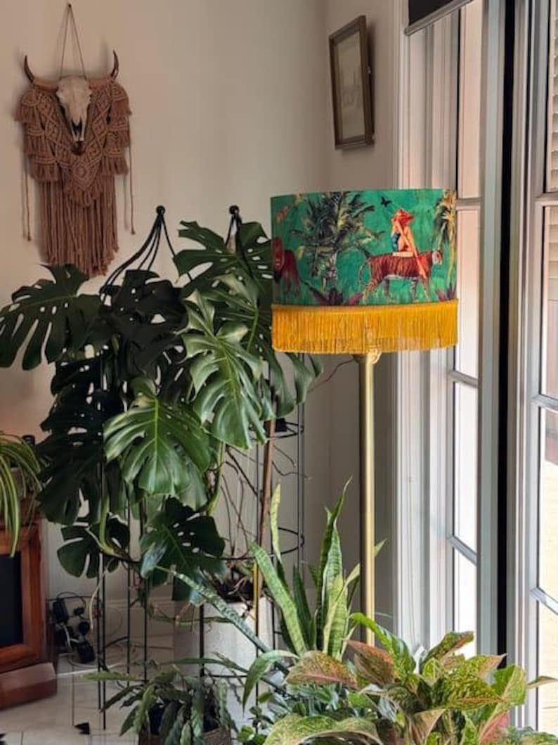 May include: A floor lamp with a green lampshade featuring a jungle scene with a tiger and a woman. The lampshade has a yellow fringe at the bottom. The lamp is gold-colored and stands among various houseplants.