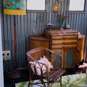 May include: A room with vintage furniture, including a wooden cabinet, a rattan chair with a pink pillow, and a floor lamp with a teal lampshade featuring a lion design. A green and white leaf-patterned rug is on the floor.