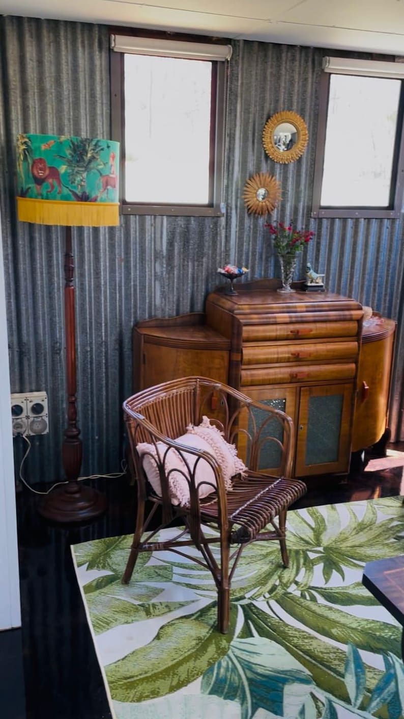 May include: A room with vintage furniture, including a wooden cabinet, a rattan chair with a pink pillow, and a floor lamp with a teal lampshade featuring a lion design. A green and white leaf-patterned rug is on the floor.