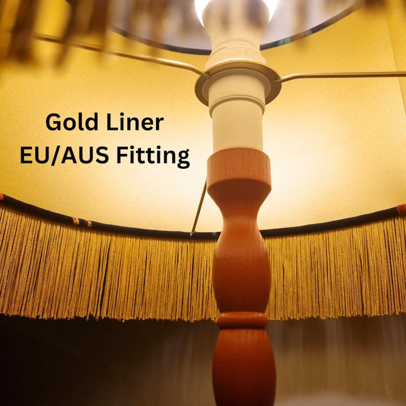 May include: A close-up of a lamp with a gold-colored shade and a brown wooden base. The shade has a gold liner and a fringe trim. The text "Gold Liner EU/AUS Fitting" is visible on the shade.