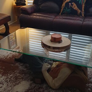 May include: A glass coffee table with a brown and white cowhide rug underneath. A wooden coaster with a red and white ceramic object on top sits on the table. The table is in front of a brown leather sofa with a pillow featuring a green, black, and gold pattern.