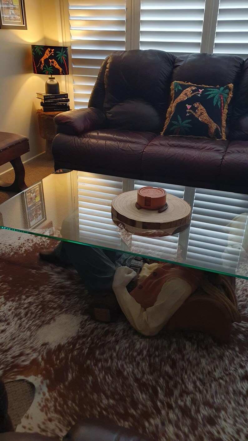 May include: A glass coffee table with a brown and white cowhide rug underneath. A wooden coaster with a red and white ceramic object on top sits on the table. The table is in front of a brown leather sofa with a pillow featuring a green, black, and gold pattern.