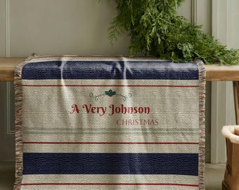 May include: A rectangular throw blanket with a cream background and horizontal stripes in navy blue, red, and green. The text "A Very Johnson CHRISTMAS" is displayed in red. The blanket is fringed at the edges.