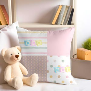 May include: A pink and white throw pillow with a patchwork design featuring stripes, polka dots, and blocks with the letters A, B, and C. The pillow is decorated with pastel colors and a watercolor effect.
