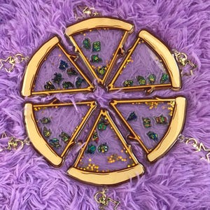 Op de afbeelding: Zes sleutelhangers in de vorm van pizzapunten met goudkleurige korsten en transparante centra. Elke sleutelhanger heeft miniatuur stripfiguren en goudkleurige sterren-glitter. De sleutelhangers zijn gerangschikt op een paars, fuzzy oppervlak.