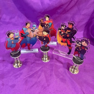 May include: A collection of five acrylic character standees featuring various superhero duos in chibi art style. Three standees are mounted on silver metal springs, while two are on clear acrylic bases. The figures depict characters in colorful costumes.