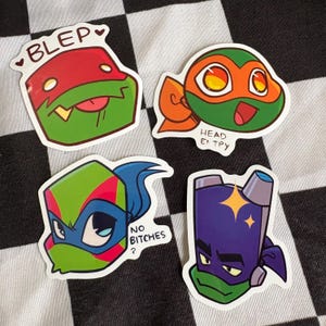 May include: Four colourful stickers featuring cartoon turtle characters. One sticker has the word "BLEP" above the turtle's head. Another sticker has the words "HEAD EMPTY". A third sticker has the words "NO BITCHES?". The stickers are on a black and white checkered surface.