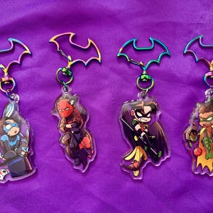 Peut inclure: Quatre porte-clés en acrylique représentant des personnages de DC Comics. Chaque porte-clés a un symbole de chauve-souris coloré en haut et un design de personnage en dessous. Les personnages incluent Nightwing, Red Hood, Robin et une autre variante de Robin.