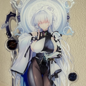 May include: A clear acrylic standee featuring a stylized anime character with long white hair and a black and white outfit. The character is set against a light blue and white circular backdrop with intricate designs. The standee has a glossy finish.