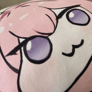 Könnte beinhalten: Nahaufnahme eines rosa-weißen Plüschtiers mit einem Comic-Gesicht. Das Spielzeug hat große, schwarz umrandete Augen mit lila Pupillen und einen kleinen, schwarz umrandeten Mund.