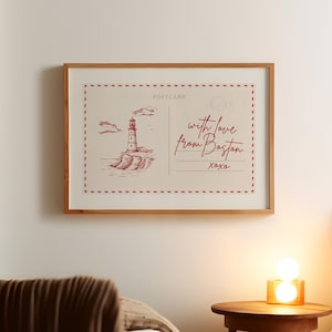 Op de afbeelding: Ingelijste ansichtkaartkunst met een rood-witte vuurtorenillustratie met de tekst "with love from Boston xoxo". Het kunstwerk zit in een houten lijst en hangt aan een witte muur.