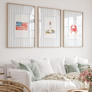 Peut inclure: Trois impressions encadrées à l'aquarelle sur le thème nautique sont accrochées au-dessus d'un canapé blanc. Les impressions représentent un drapeau américain, un phare et un crabe rouge. Les cadres ont un fond à rayures bleu clair.