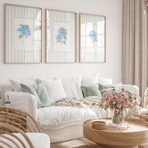 Puede incluir: Tres cuadros con estampados botánicos de hortensias azules cuelgan sobre un sofá blanco con cojines verdes y blancos. Una mesa de centro de madera contiene un jarrón con flores rosas y una cesta tejida. La habitación tiene una paleta de colores neutros.