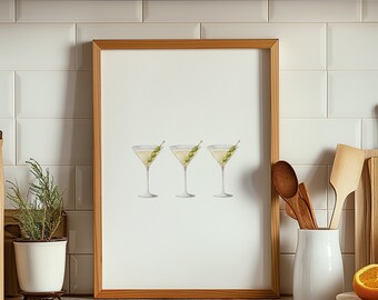 May include: A framed print featuring three watercolor martini glasses, each garnished with green olives. The artwork is set against a white background and framed in light brown wood. The print is displayed on a kitchen counter.