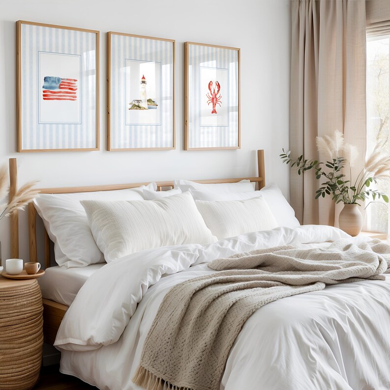 Puede incluir: Tres impresiones enmarcadas en acuarela con temas n&aacute;uticos cuelgan sobre una cama con ropa de cama blanca. Las impresiones presentan una bandera estadounidense, un faro y una langosta roja. Los marcos son de madera clara y el fondo es un patr&oacute;n de rayas azules y blancas.