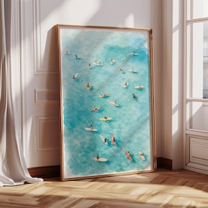 May include: A framed art print featuring a watercolor painting of surfers in the ocean. The artwork depicts numerous people on surfboards and kayaks in turquoise water. The frame is gold-colored, and the print is displayed against a white wall and wooden floor.