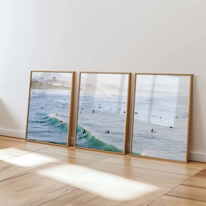 May include: Three framed prints depicting a coastal scene with surfers. The ocean is a vibrant blue-green, with many surfers. The frames are light wood, creating a serene aesthetic. The prints capture the essence of surfing and the beauty of the coast.