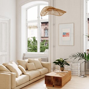 May include: A living room with a beige sofa, wooden coffee table, and a framed artwork of an American flag. A woven, saucer-shaped light fixture hangs from the ceiling. A large window offers a view of the outside.